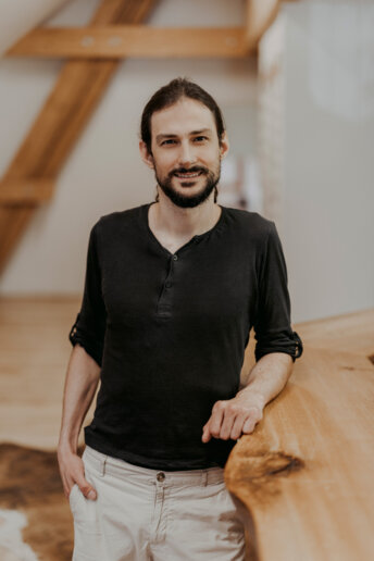 Lächelnder Mann mit zurückgebundenem Haar und Bart, schwarzem Henley-Shirt und heller Hose, lehnt an einer massiven Holzkante in einem hellen Raum mit Holztreppe im Hintergrund.
